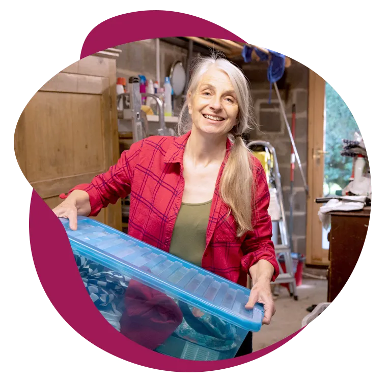A smiling woman with long gray hair carries a clear storage bin through her garage.